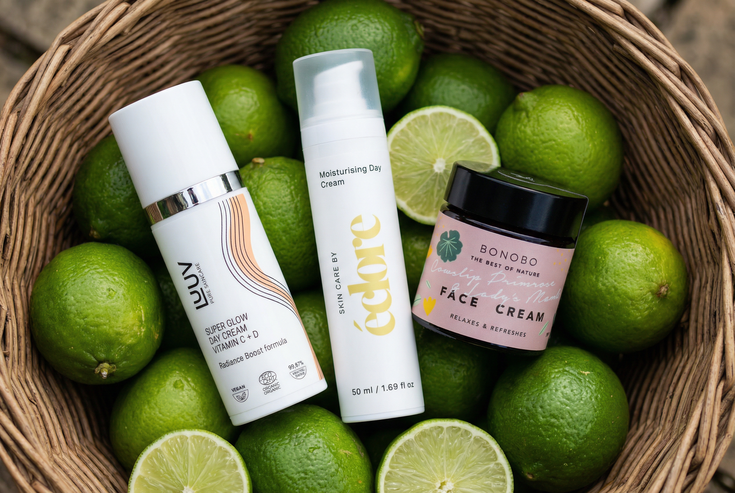 Tre naturliga ansiktskrämer i en flätad korg fylld med limefrukter – LUUV Super Glow Day Cream Vitamin C+D, éclore Moisturising Day Cream och BONOBO Face Cream. Ekologiska och veganska dagkrämer för lyster och återfuktning.