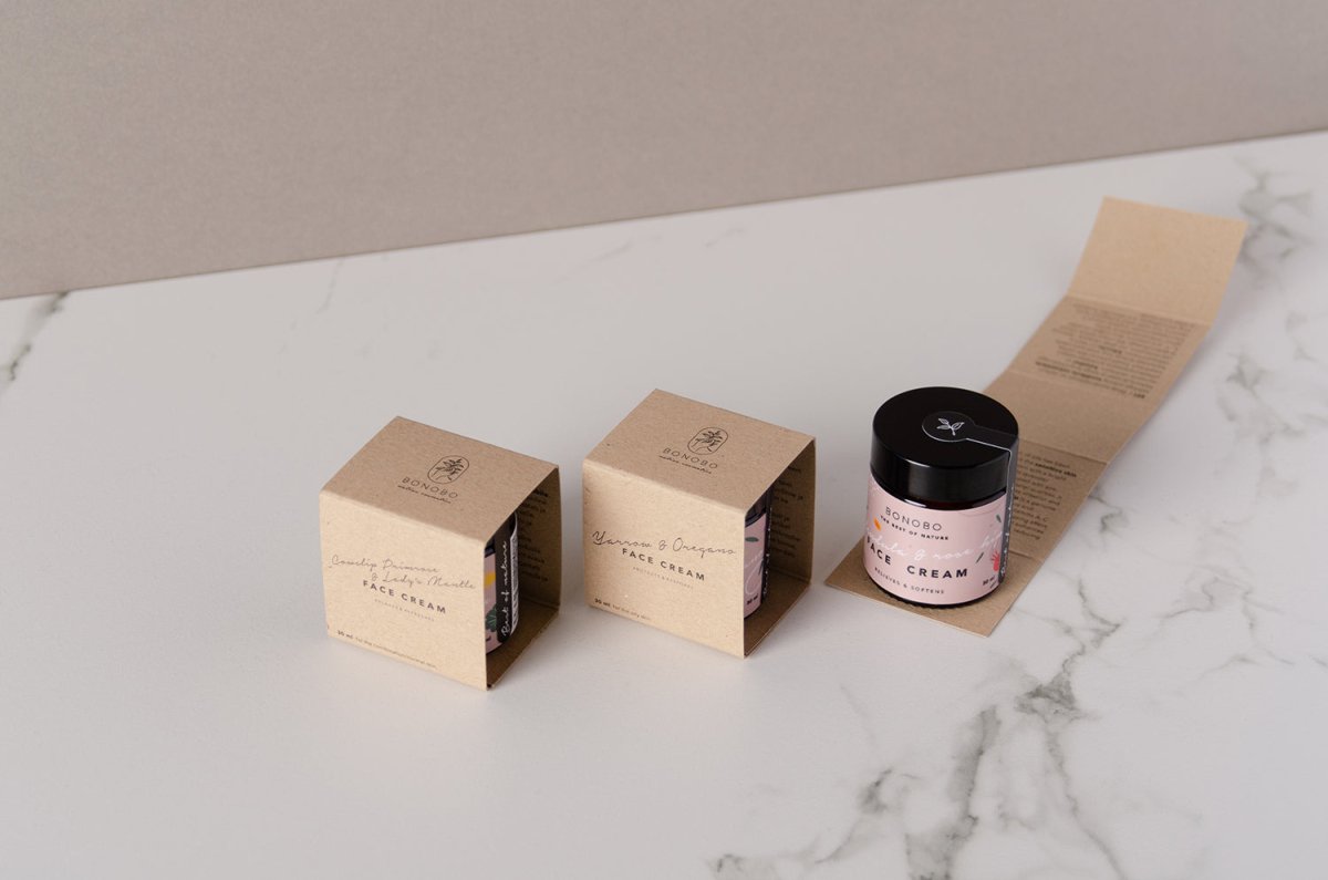 Naturlig ansiktskräm med ringblomma och nypon i rosa burk med svart lock, bredvid beige förpackningskartonger