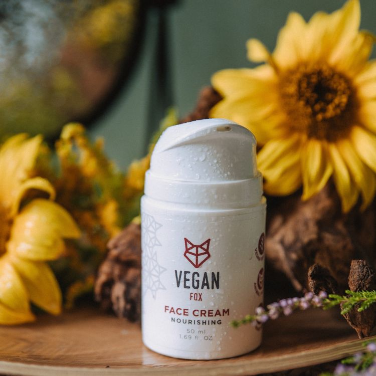 Vit pumpflaska med Vegan Fox Face Cream står på träunderlag omgiven av gula blommor och gröna växter