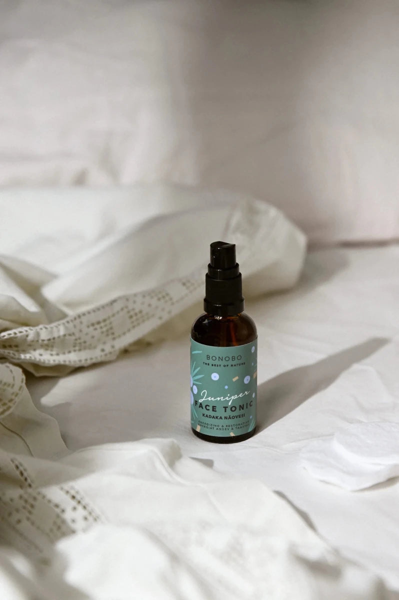 Brun glasflaska med svart spraytopp innehåller Bonobo Juniper Face Tonic, omgiven av vita textilier på ljus bakgrund