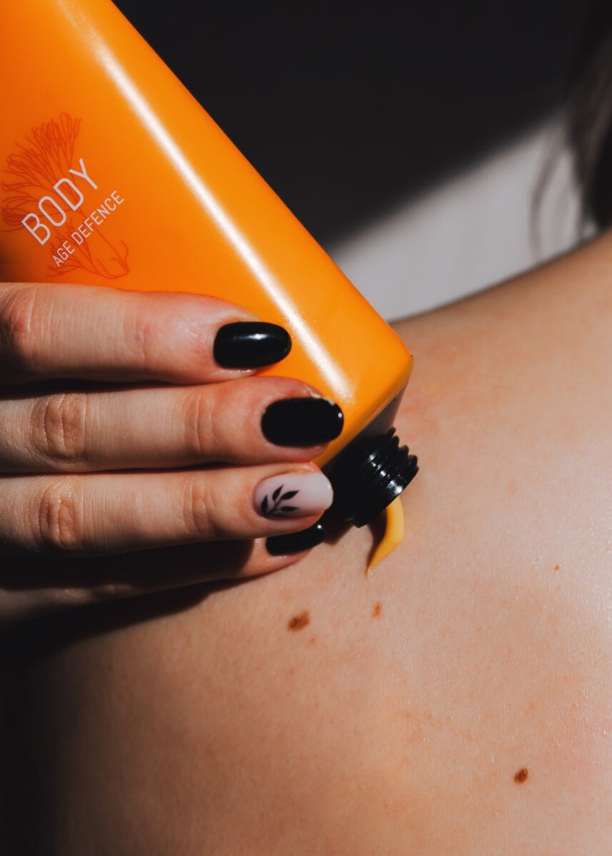 Orange tube med Age Defence BODY Cream appliceras på hud med händer med svart nagellack
