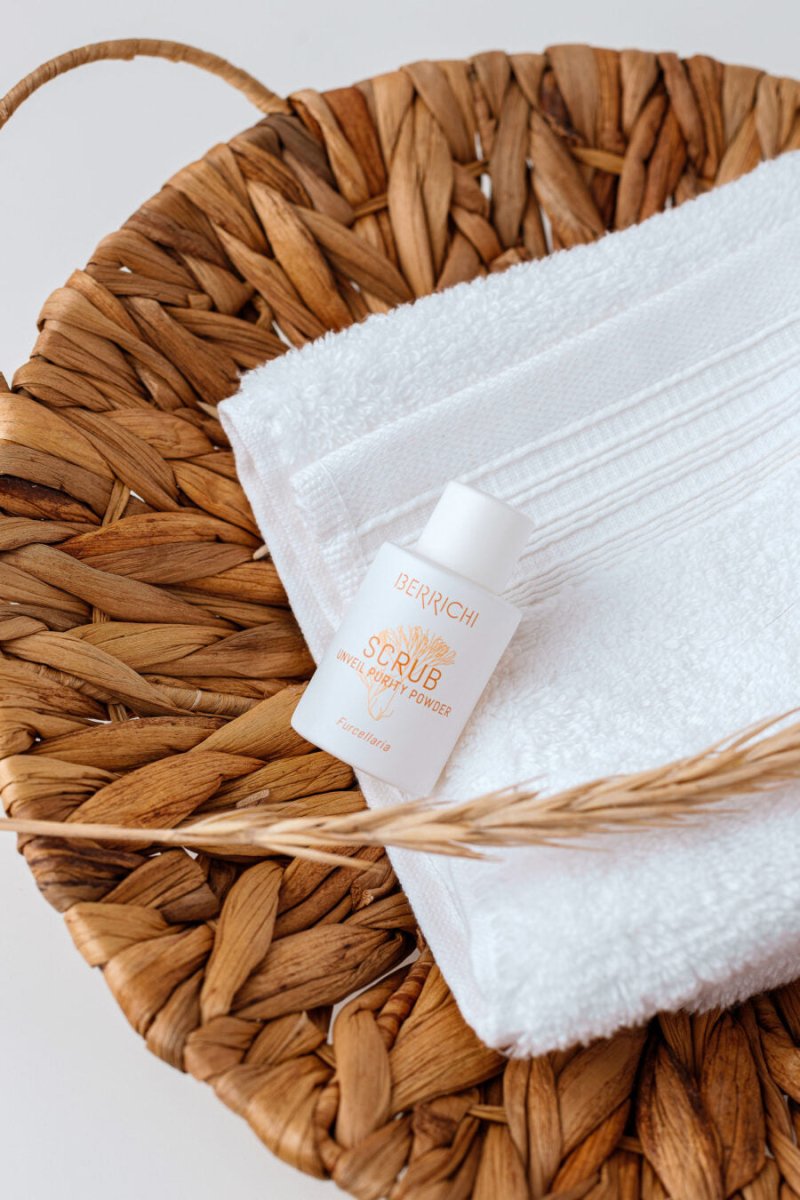 Reveal Purity Puderskrubb i vit förpackning med orange text ligger på fluffig handduk i korgkorg med vetekorn