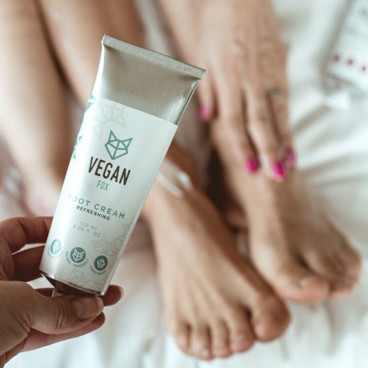 Vit tub med fotkräm 120 ml hålls i hand, med avslappnade fötter med rosa nagellack i bakgrunden på vit säng