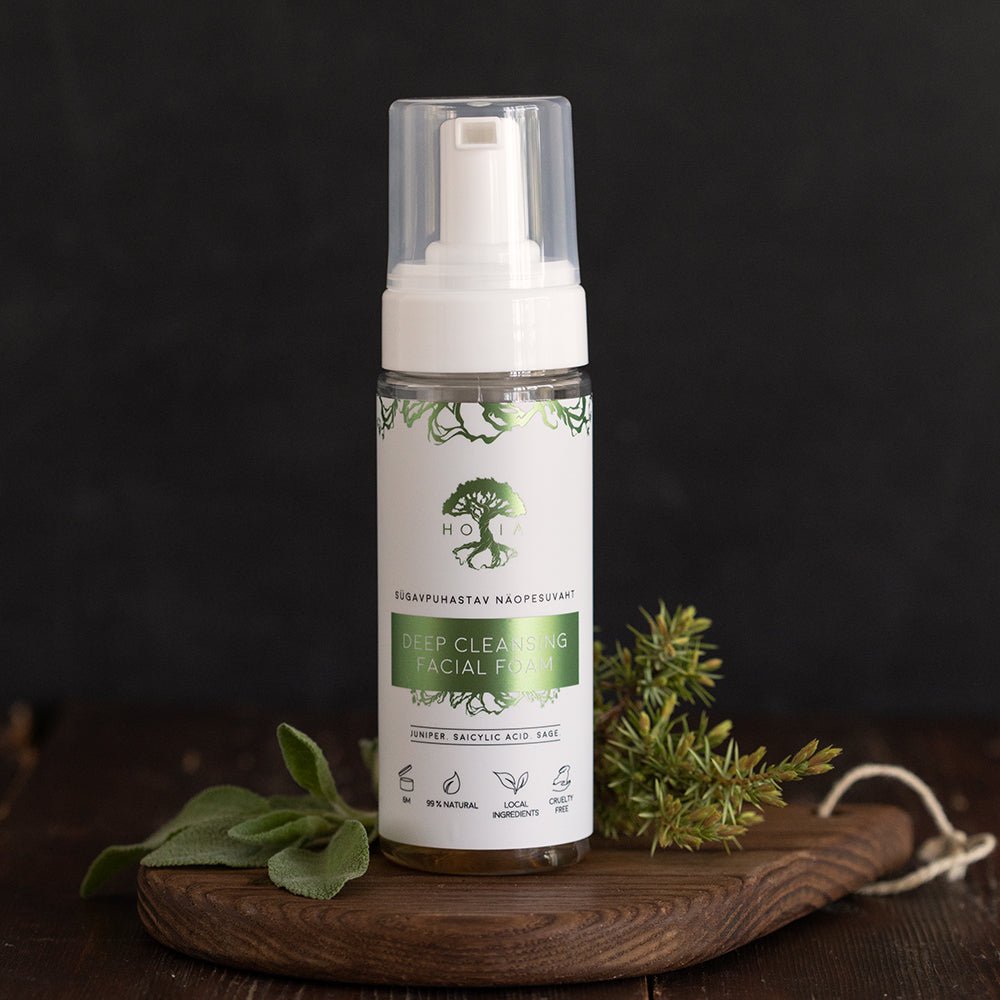 Vit sprayflaske med transparent pump og grønn etikett som viser Deep Cleansing Facial Foam på trebrett med urter