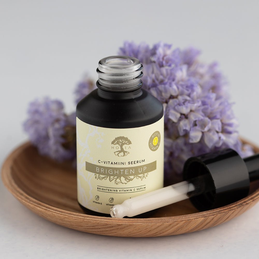 Brightening Vitamin C Serum i mörk glasflaska med pipett på träbricka, omgiven av lila blommor
