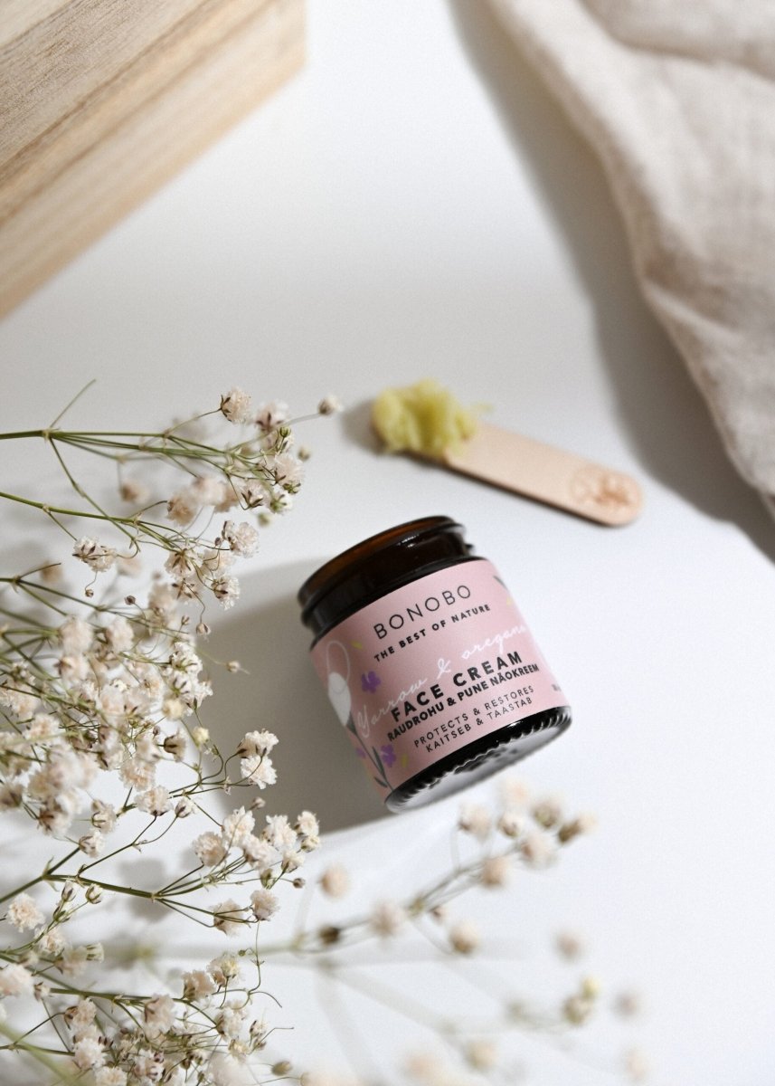 BONOBO ansiktskräm med rölleka och oregano i rosa glasburk med träspatel på vit bakgrund med torkade blommor