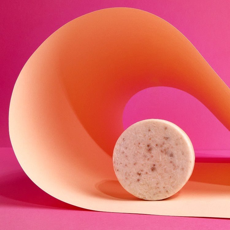 Rund rosa ansiktsrengöringsbar med Rosa Lera 50g, spräcklig textur, framför orange böjd form på rosa bakgrund