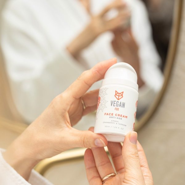 Vit burk med Anti-Age Face Cream 50ml från Vegan Fox hålls i händerna, person i vit badrock i bakgrunden
