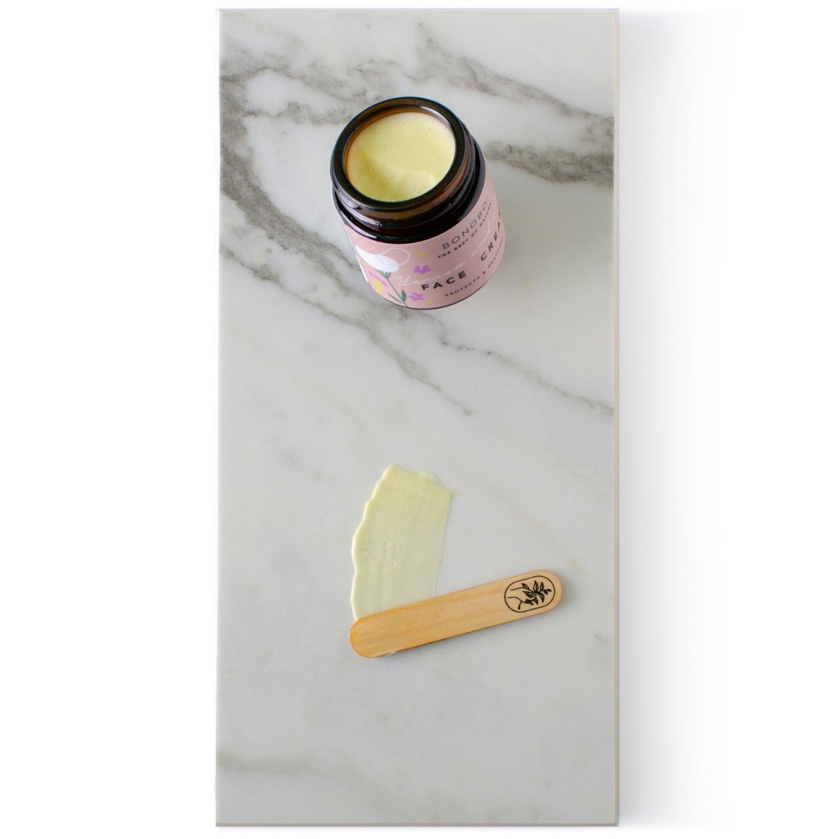 Ansiktskräm Rölleka och Oregano i rosa burk med gul kräm synlig, träsked med krämprov på marmorbakgrund