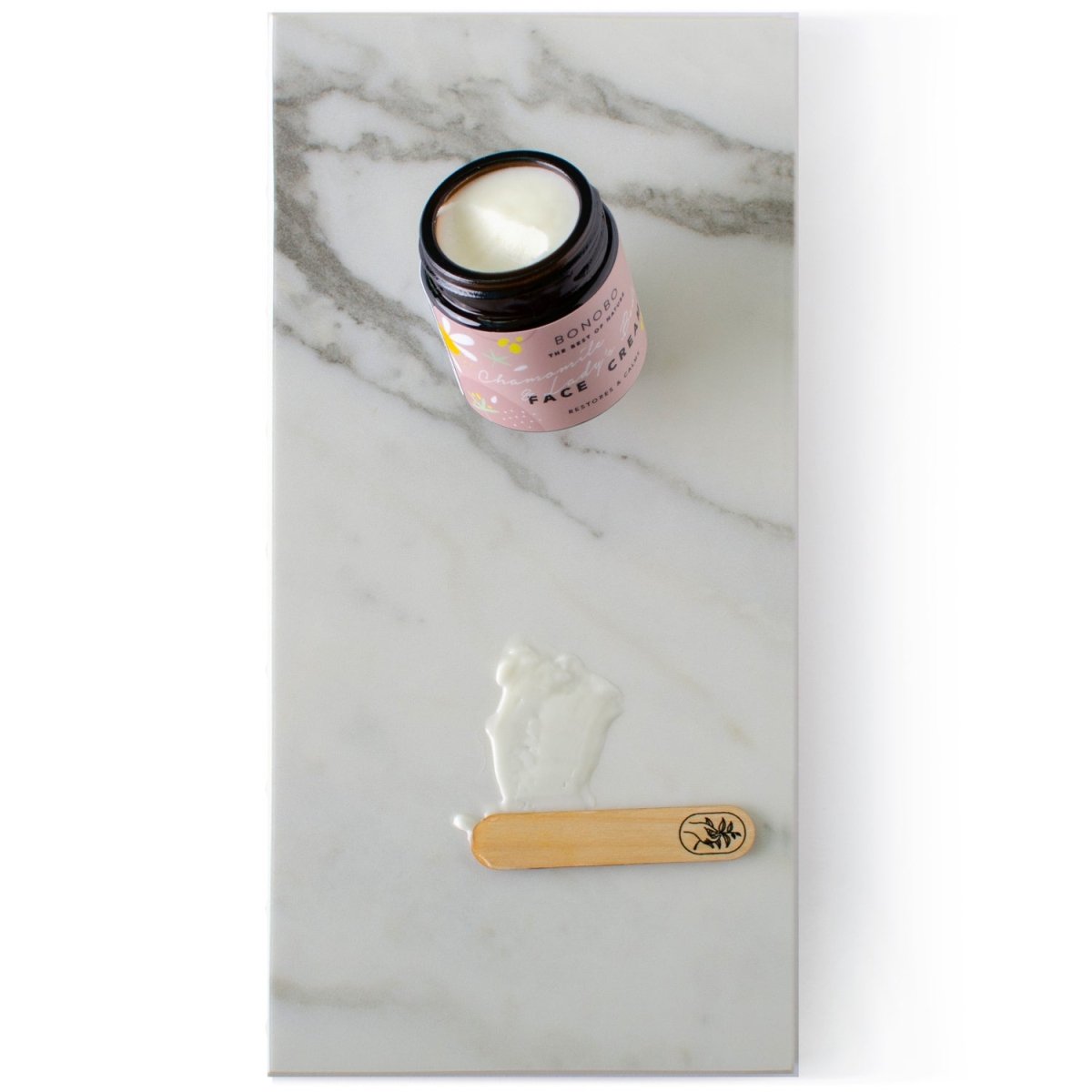 Öppen burk med ansiktskräm med kamomill och måra, rosa etikett, vit kräm synlig, träsked bredvid på marmorbakgrund