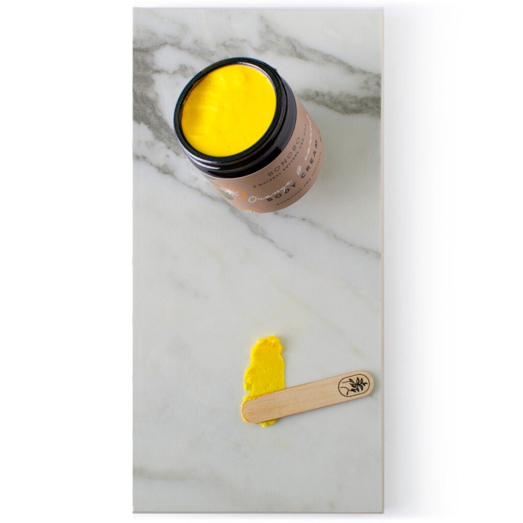 Body Cream Apelsin & Morot i öppen burk med gul kräm och träspatel med produktprov på marmorbakgrund