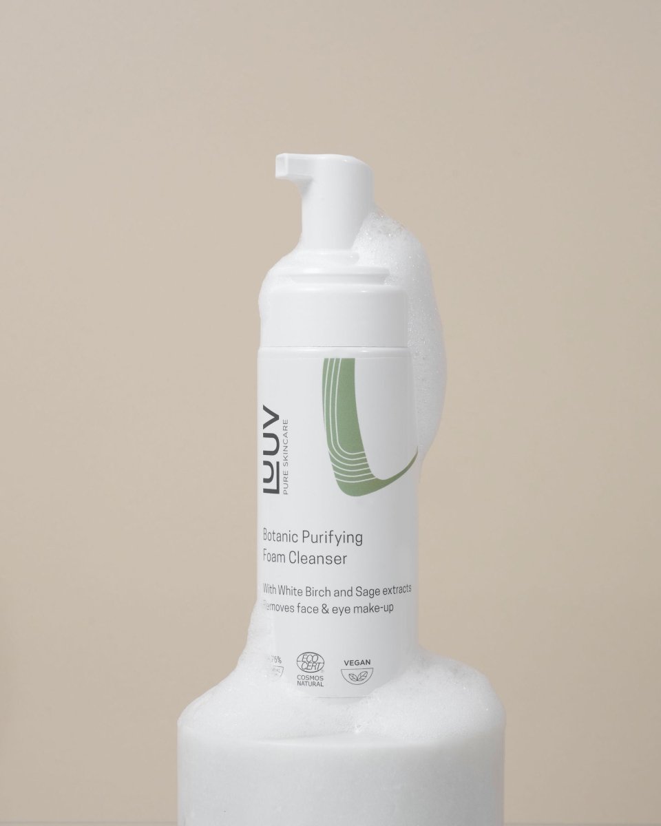 Vit flaskpump med Botanic Purifying Foam Cleanser omgiven av skummande bubblor mot beige bakgrund