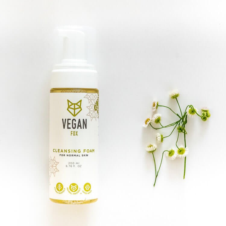 Vit flaska med gulddetaljer innehåller Vegan Fox rengöringsskum för normal hud 200 ml, bredvid små vita blommor
