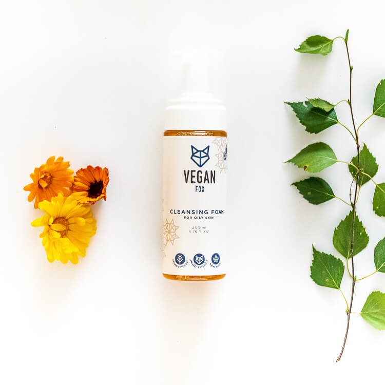 Vit cylinderformad flaska med orange detaljer innehållande rengöringsskum för fet hud, omgiven av gula blommor