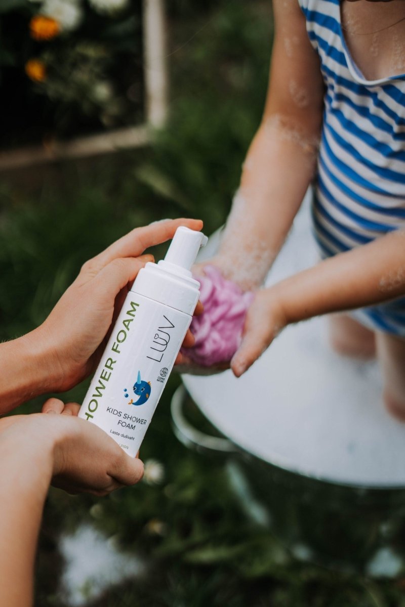 Vit sprayflaska med Duschkräm för barn Päron COSMOS hålls i hand, barn i randig tröja badar med rosa tvål i bakgrunden