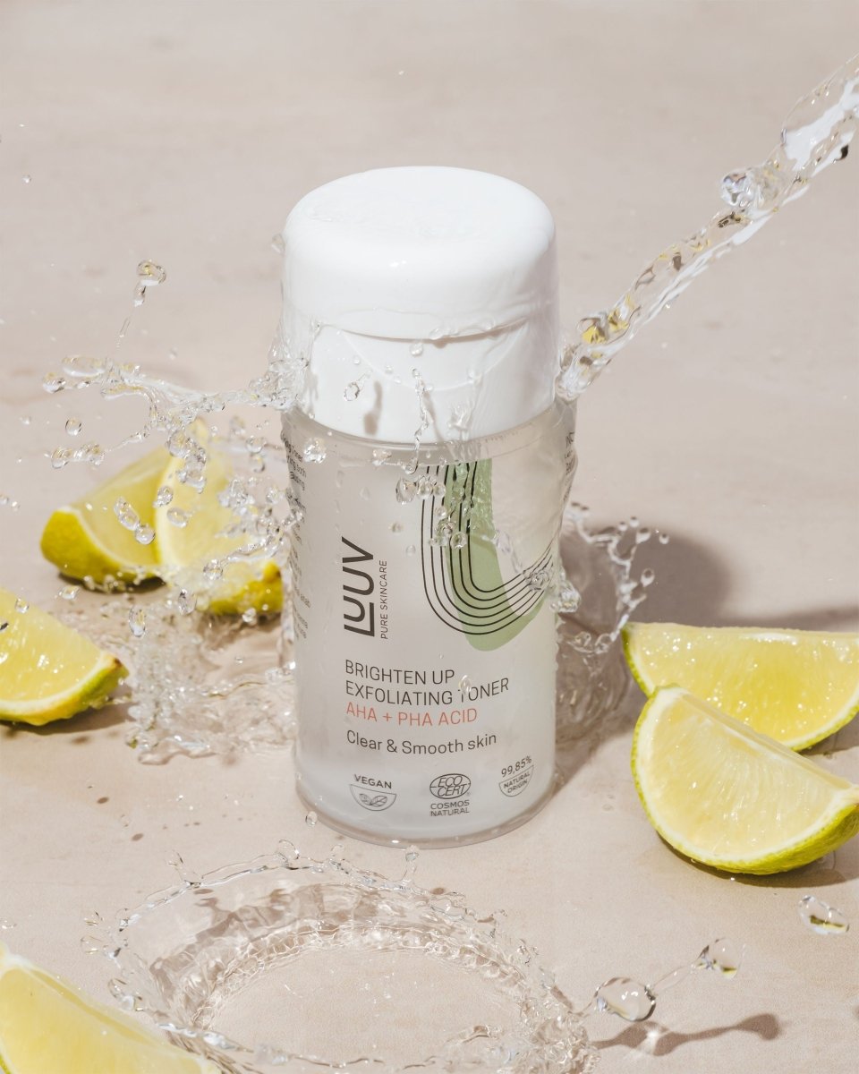 Exfolierande Ansiktstoner AHA + PHA Syror i genomskinlig flaska med vit kork, omgiven av citruskivor och vattenstänk