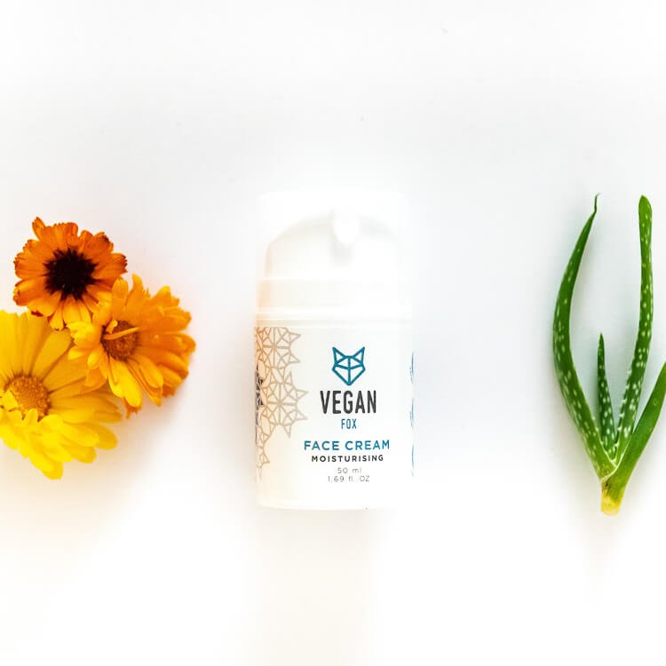 Vit burk med Vegan Fox Moisturising Face Cream 50ml omgiven av orange blommor och aloe vera-blad på vit bakgrund