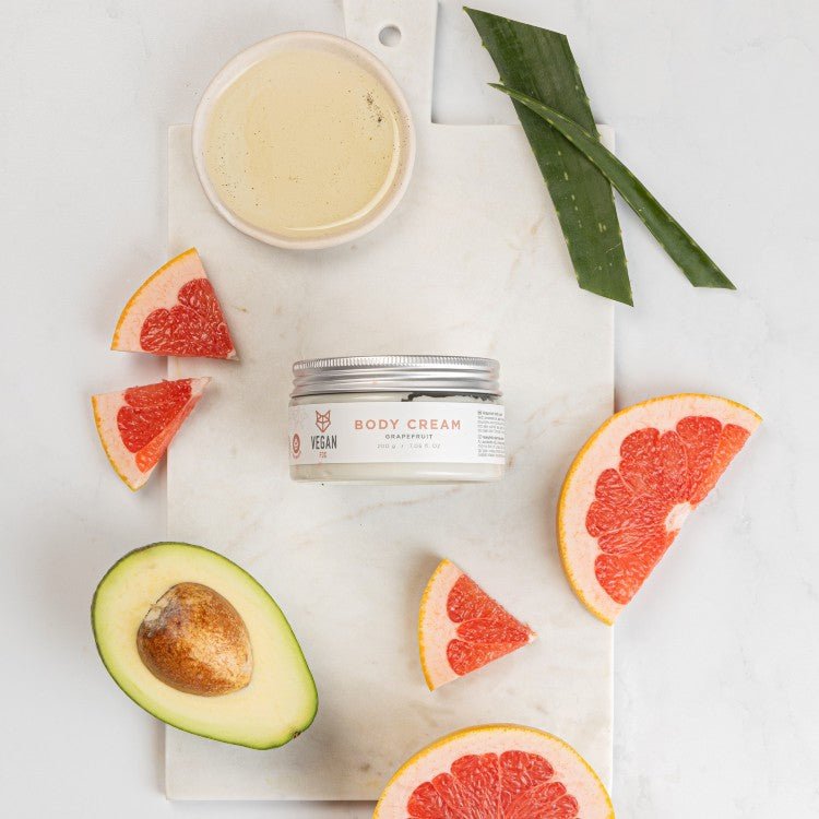 Vit burk med Grapefrukt Body Cream omgiven av grapefruktsklyftor, avokado och gröna blad på marmorbakgrund