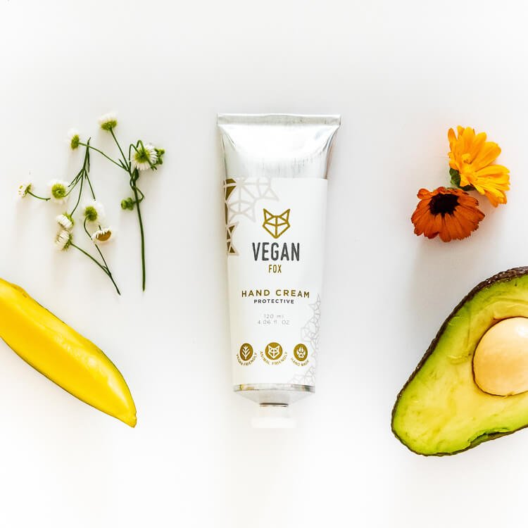 Vit tub med Vegan Fox skyddande handkräm omgiven av naturliga ingredienser som avokado, gula blommor och gröna växter
