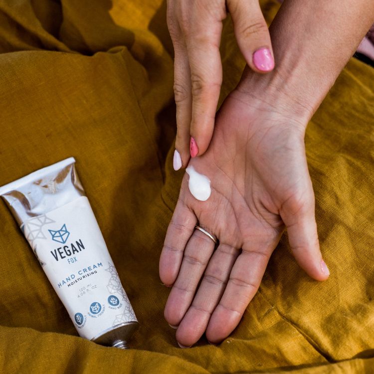 Fuktgivande handkräm med avokadoolja i vit tub, vit kräm i öppna händer med rosa naglar på gul bakgrund