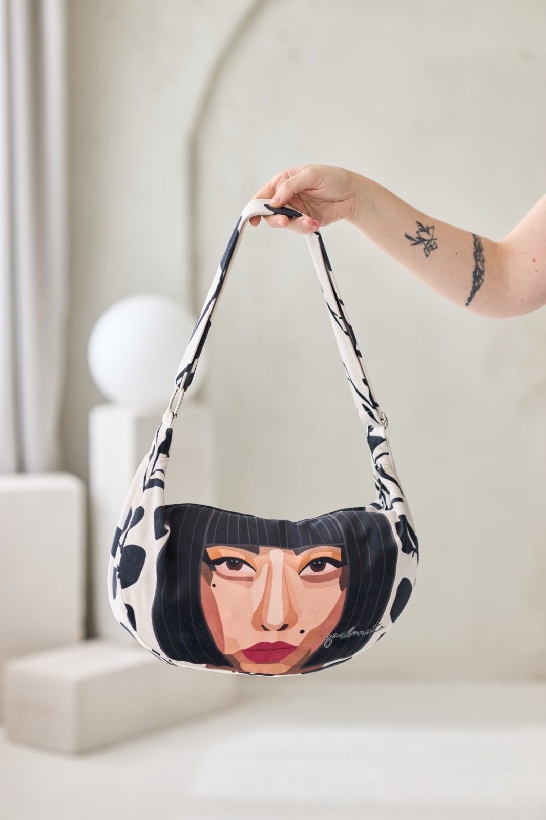 KIYOKO Hobo Handväska Velour med konstnärligt ansikte-motiv i beige och svart, hålls upp av hand med tatueringar