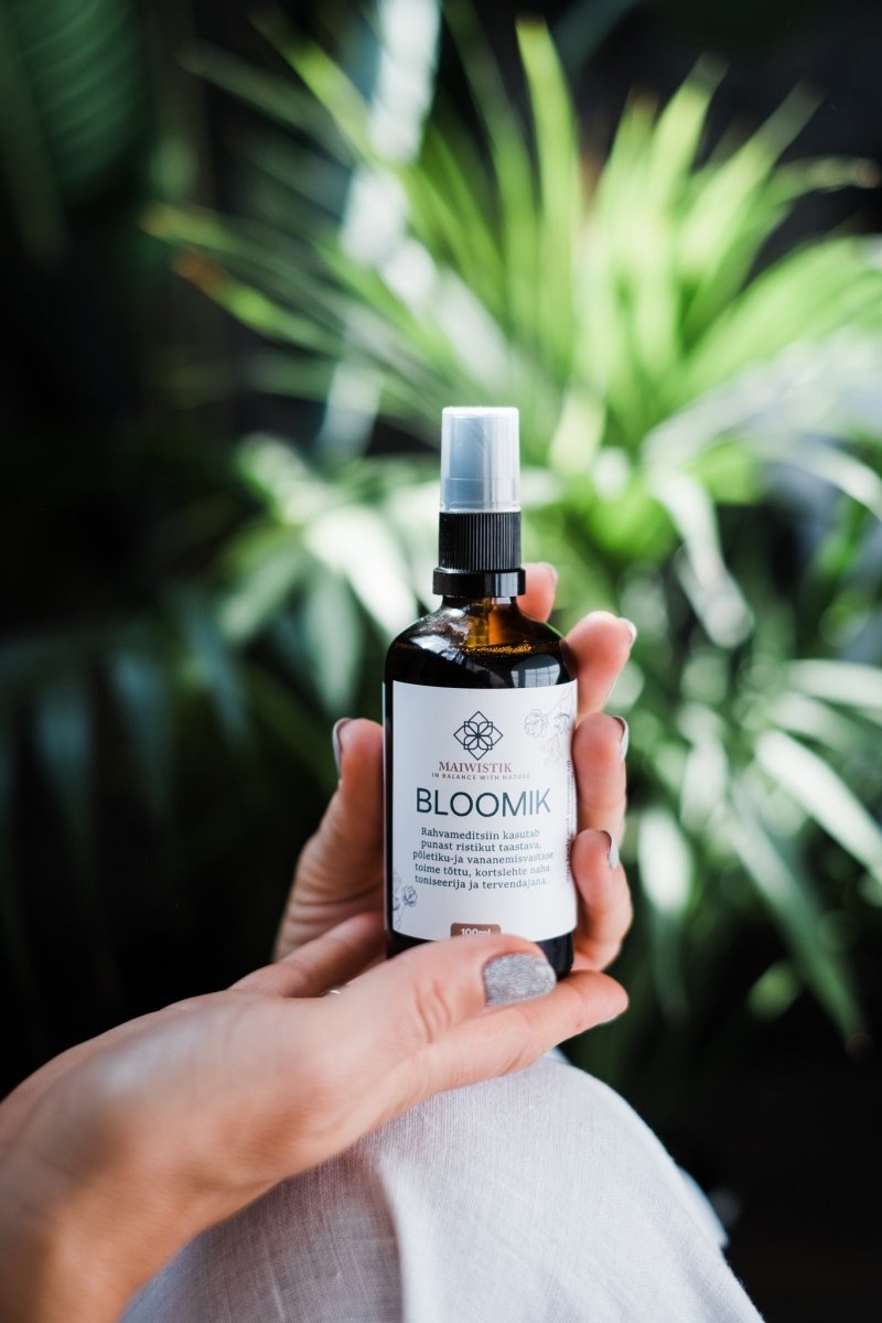 Hand håller amber glasflaska med spray och vit etikett märkt BLOOMIK, grön växtbakgrund - Kropps- och Massageolja 2i1