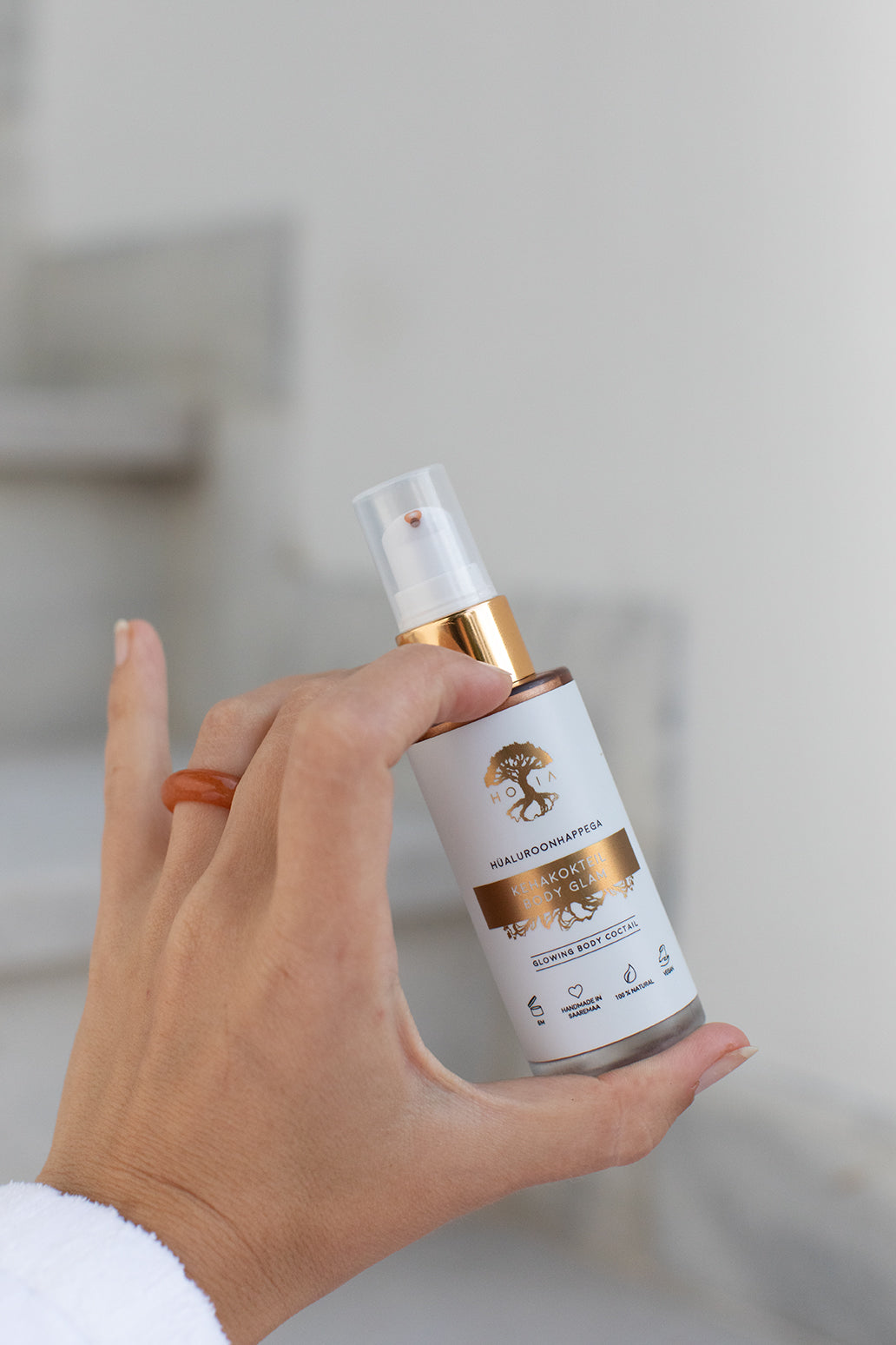 Kroppslotion Body Glam hålls i hand med spraypump, vit flaska med gulddetaljer i ljust badrum