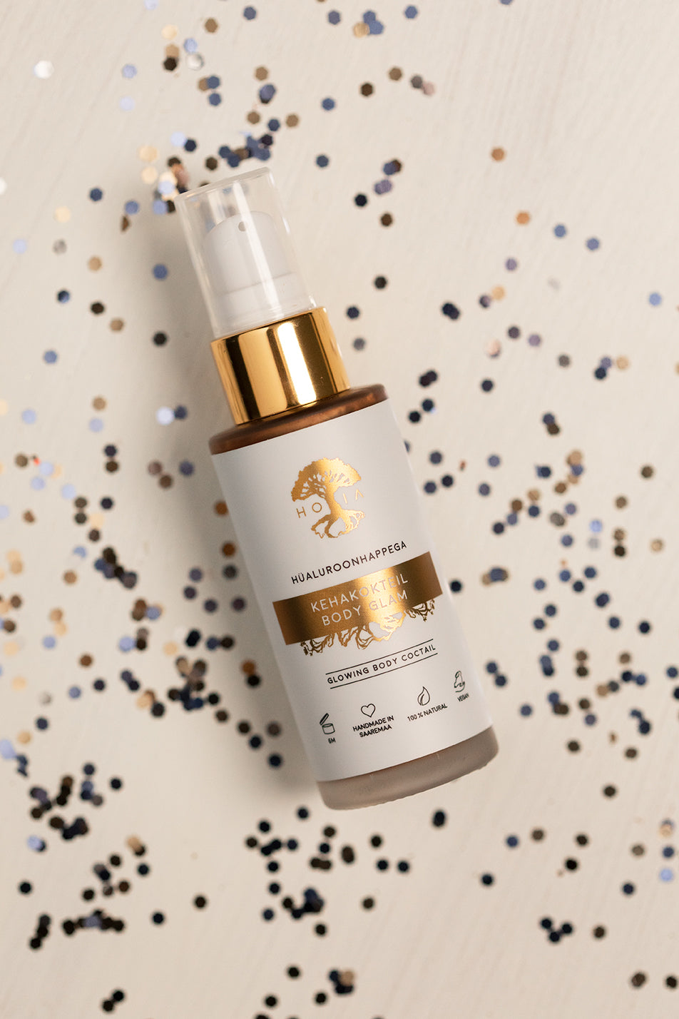 Kroppslotion Body Glam i vit pumpflaska med gulddetaljer omgiven av blått och guld konfetti på ljus bakgrund