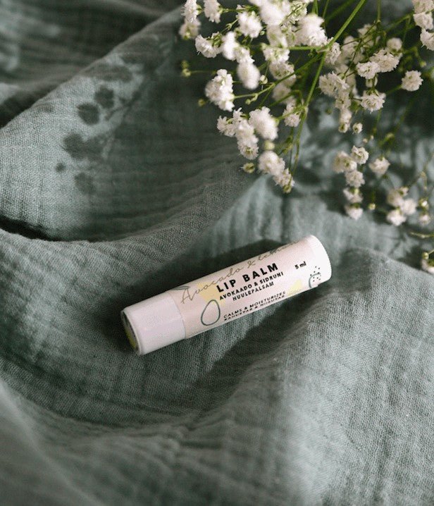 Läppbalsam Avokado & Citron i vit tub med grön text, placerad på grön linnefärg textil med vita blommor
