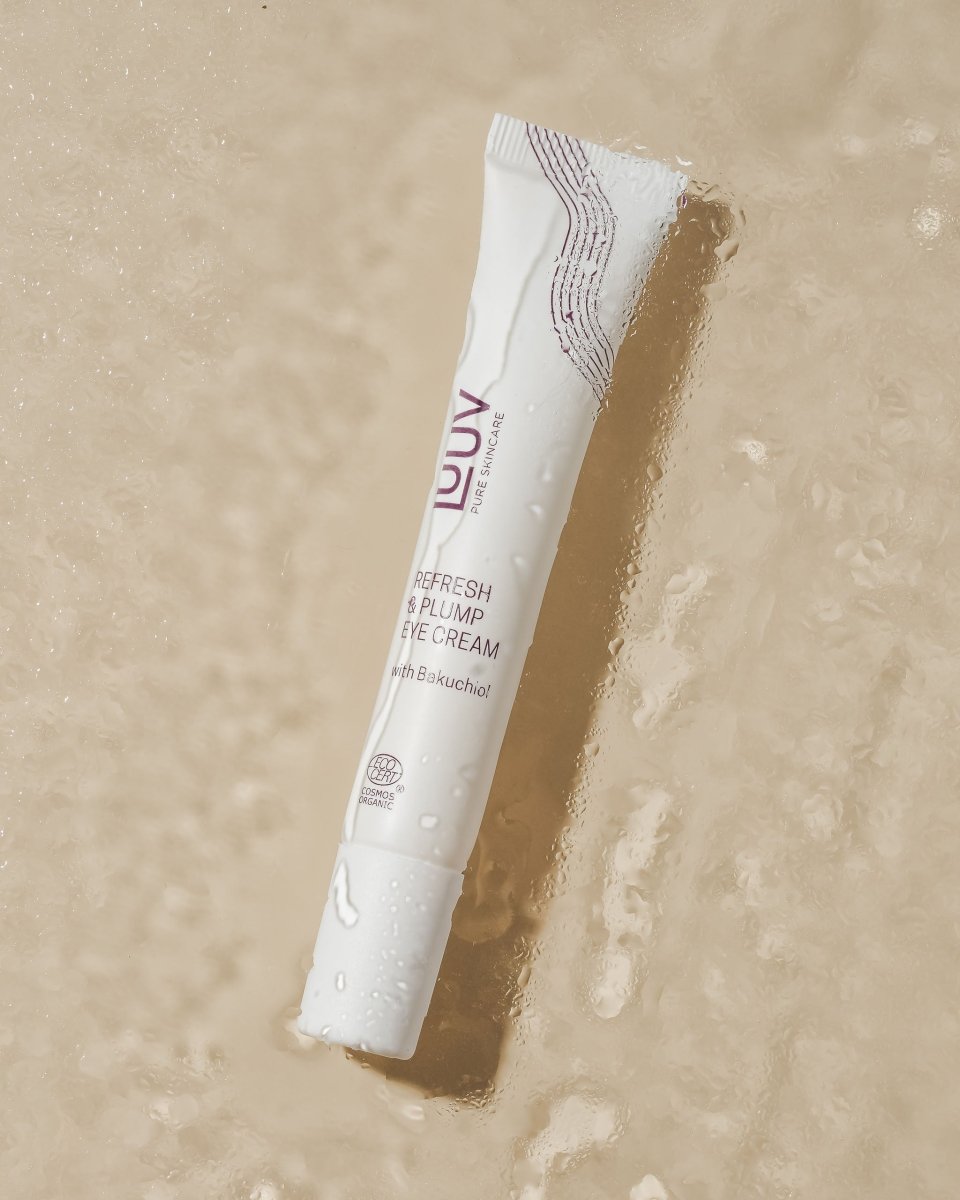 Vit tub med Refresh & Plump Eye Cream från LUUV, ligger på våt beige yta med vattendroppar och reflektioner