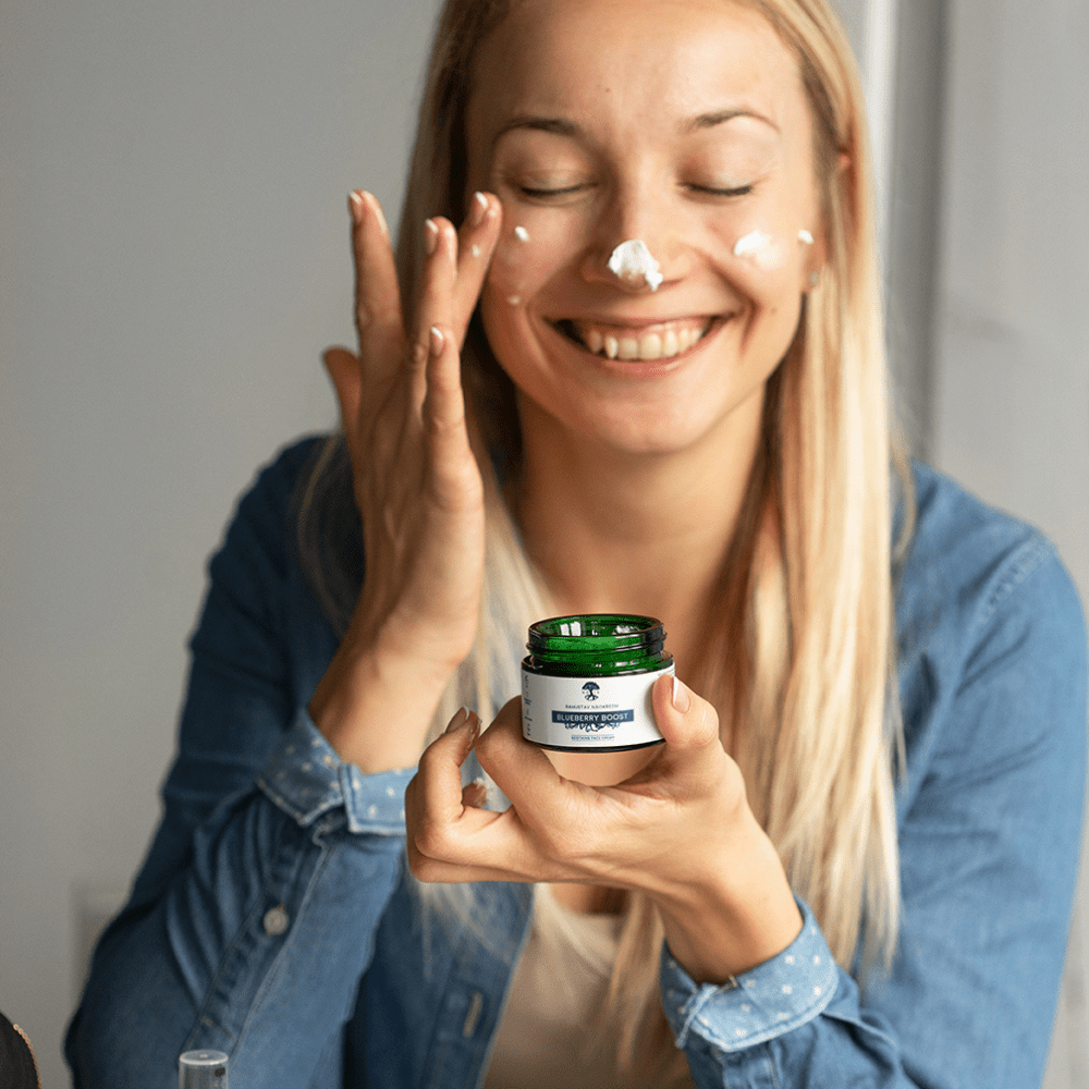 Kvinna applicerar vit kräm på ansiktet medan hon håller Lugnande Ansiktskräm Blåbär Boost 30ml i grön burk