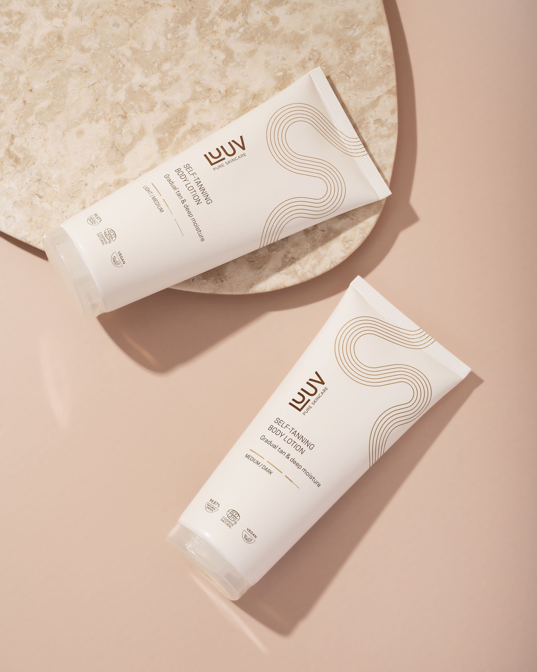 Två vita tuber med LUUV Brun Utan Sol Body Lotion, elegant design med bruna vågor, placerade på beige yta vid marmorbricka