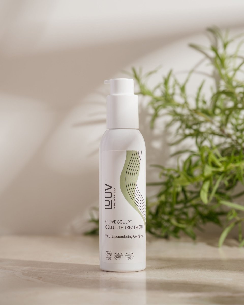 LUUV Curve Sculpt Cellulite Treatment i vit flaska med pump, stående på ljus träyta bredvid grön växt