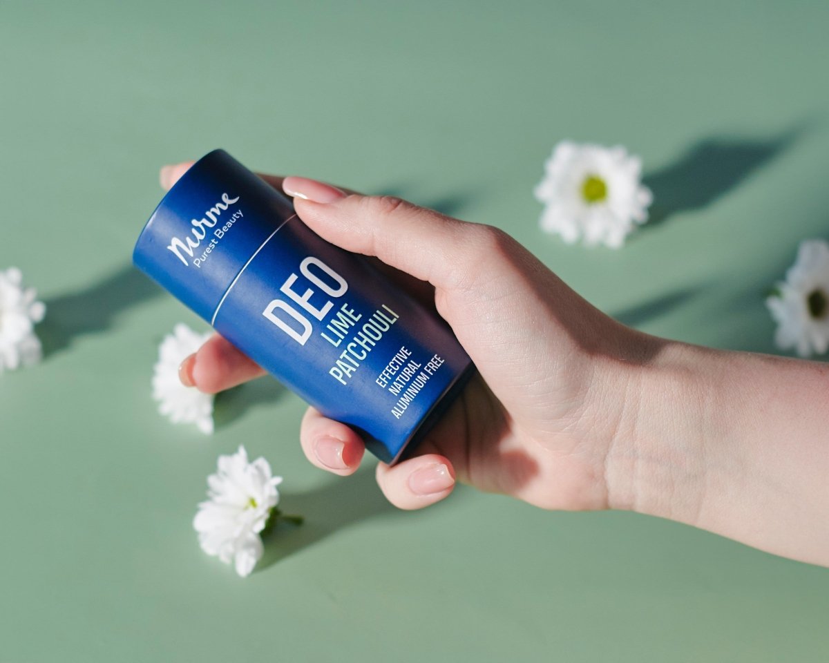 Nurme naturlig deodorant i blå förpackning med vit text hålls i hand, omgiven av vita blommor på grön bakgrund