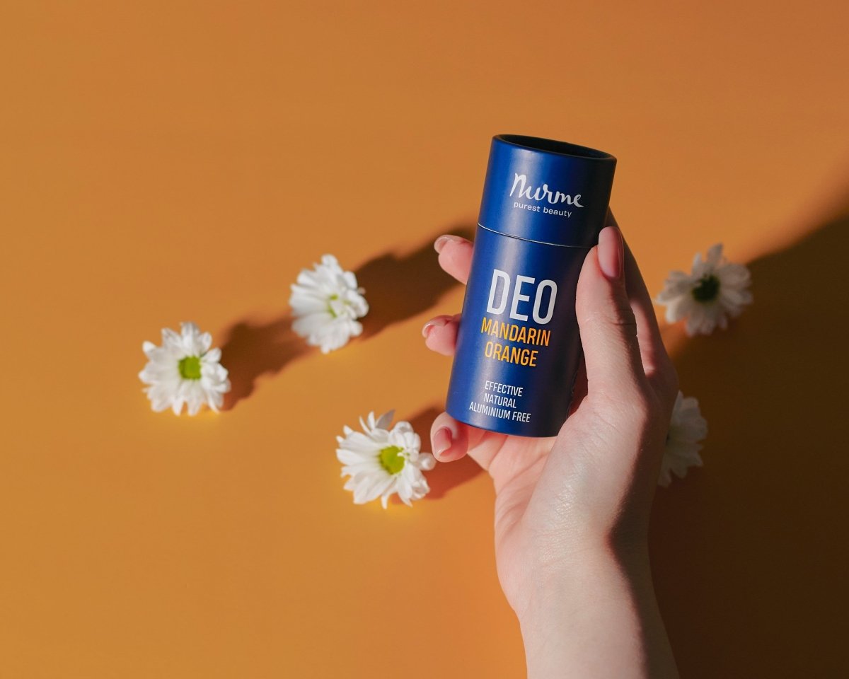 Nurme naturlig deodorant mandarin apelsin i blå cylinderförpackning hålls i hand mot orange bakgrund med vita blommor