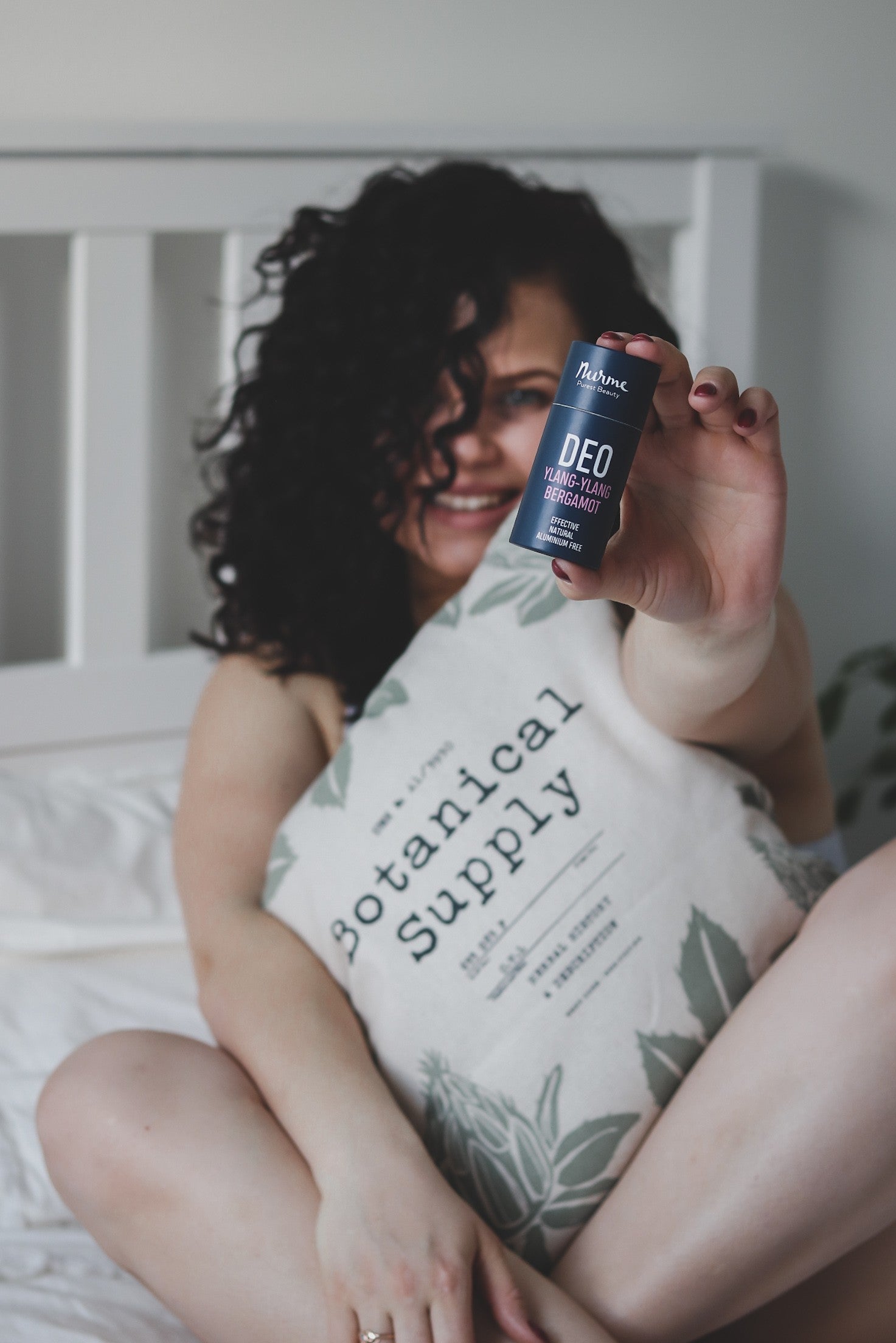 Naturlig Deodorant Ylang-Ylang & Bergamott i blå förpackning hålls upp av leende kvinna i säng