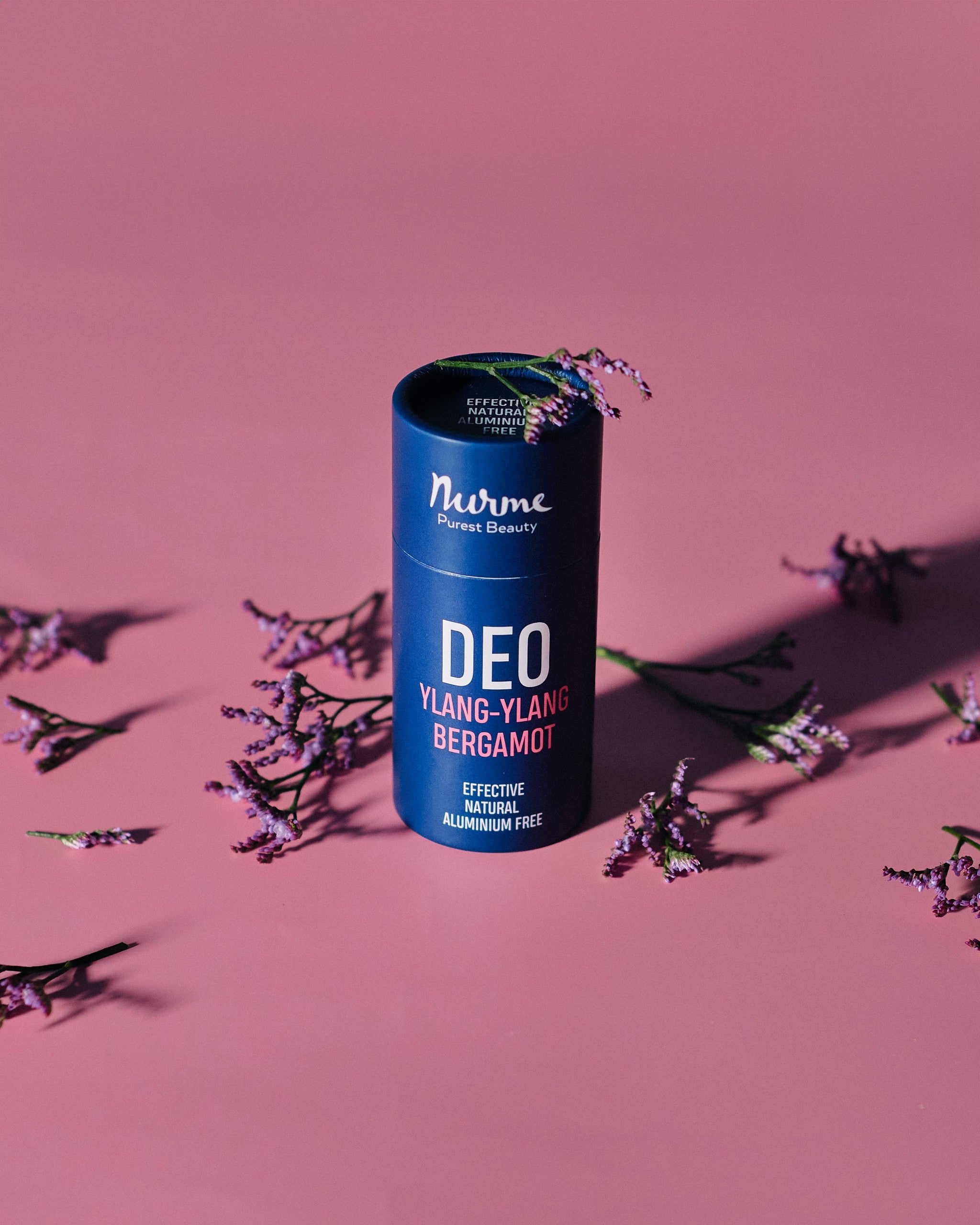 Blå cylinderformad deodorant från Nurme med text DEO YLANG-YLANG BERGAMOT, omgiven av lila lavendelkvistar på rosa bakgrund