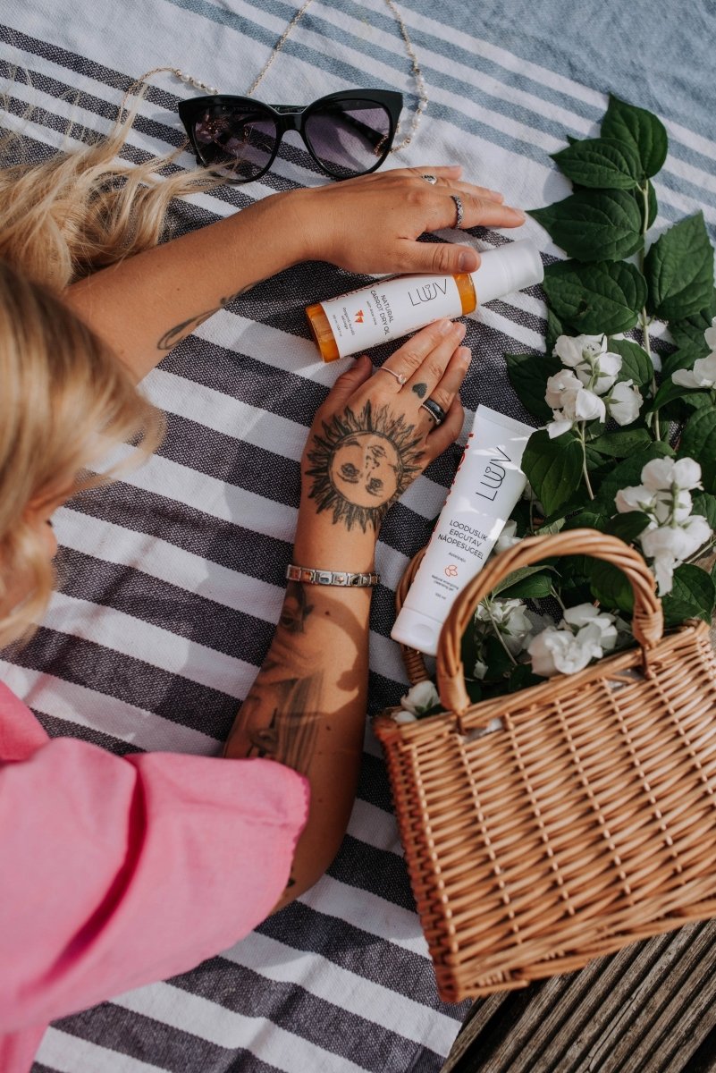 Naturlig Carrot Dry Oil med Aloe Vera i vit tub med orange detaljer hålls av händer med soltatuering på randig filt