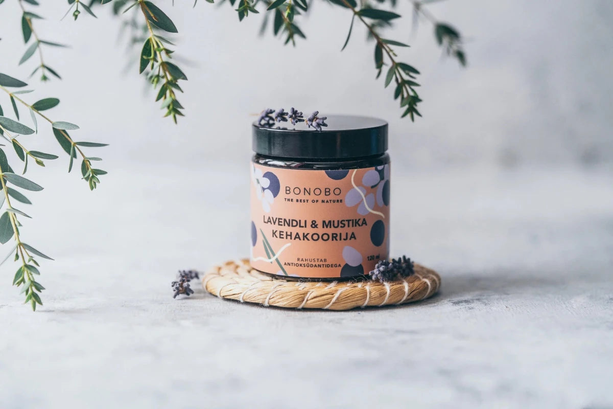 BONOBO Body Scrub med blåbär och lavendel i glasburk med svart lock, omgiven av gröna kvistar