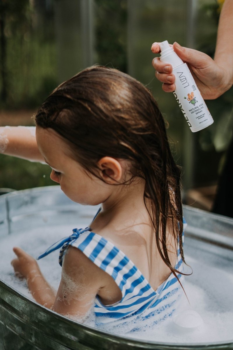 Naturligt detangler spray appliceras på barn i badkar med blårandig baddräkt, vit sprayflaska i hand