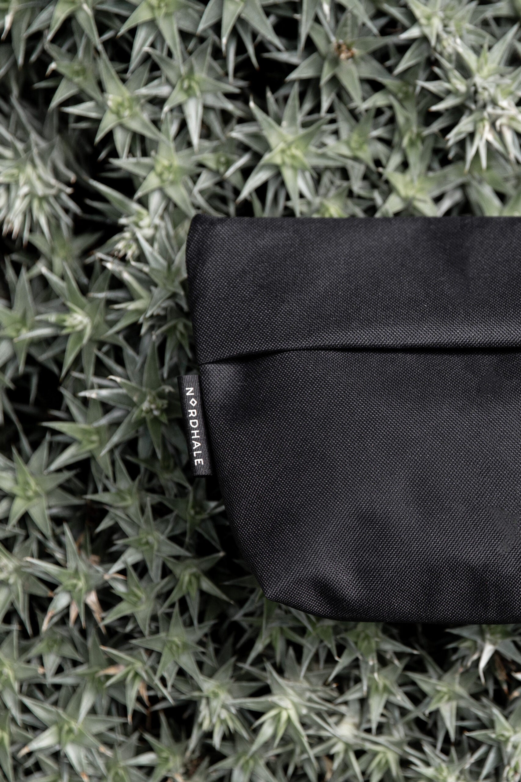 NES Toiletry Bag från Nordhale - svart premium sminkväska i textil med diskret logotyp, fotograferad mot naturlig grön växtbakgrund