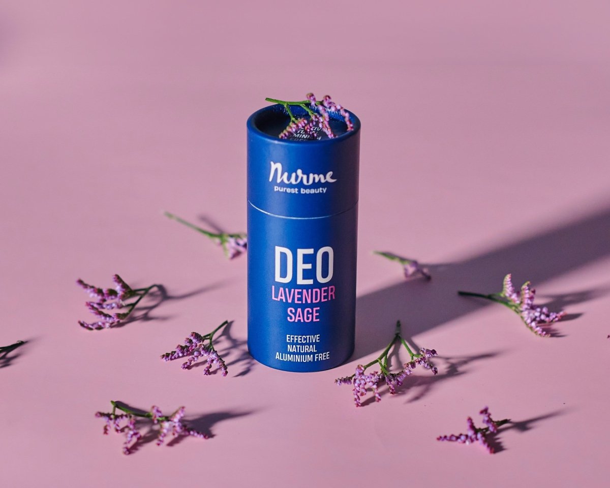 Nurme naturlig deodorant i blå cylinderförpackning med lavendel och salvia, omgiven av lila lavendelblommor på rosa bakgrund