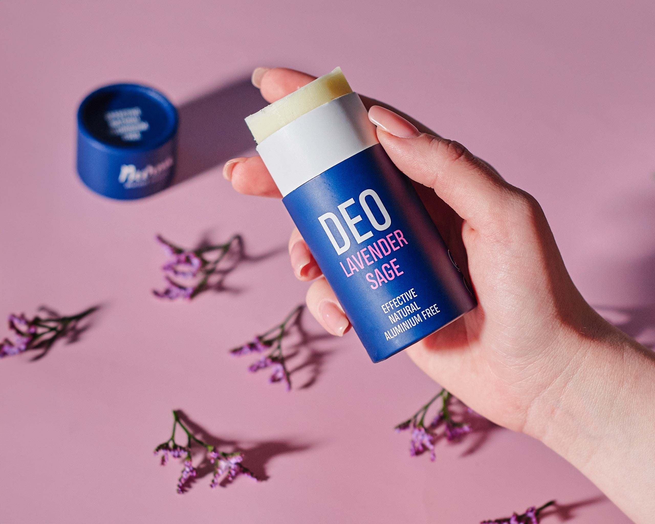 Hand håller upp öppnad Nurme naturlig deodorant lavendel och salvia med synlig stickprodukt, lavendelblommor och öppnat lock i bakgrunden