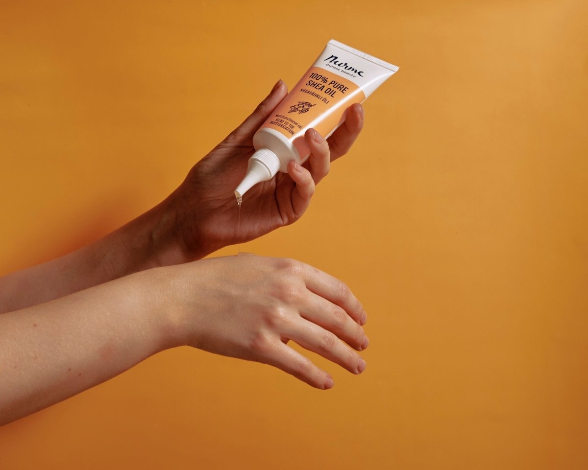 Nurme Organic Shea Oil i vit tub med orange detaljer hålls i hand mot orange bakgrund, produkten pressas ut