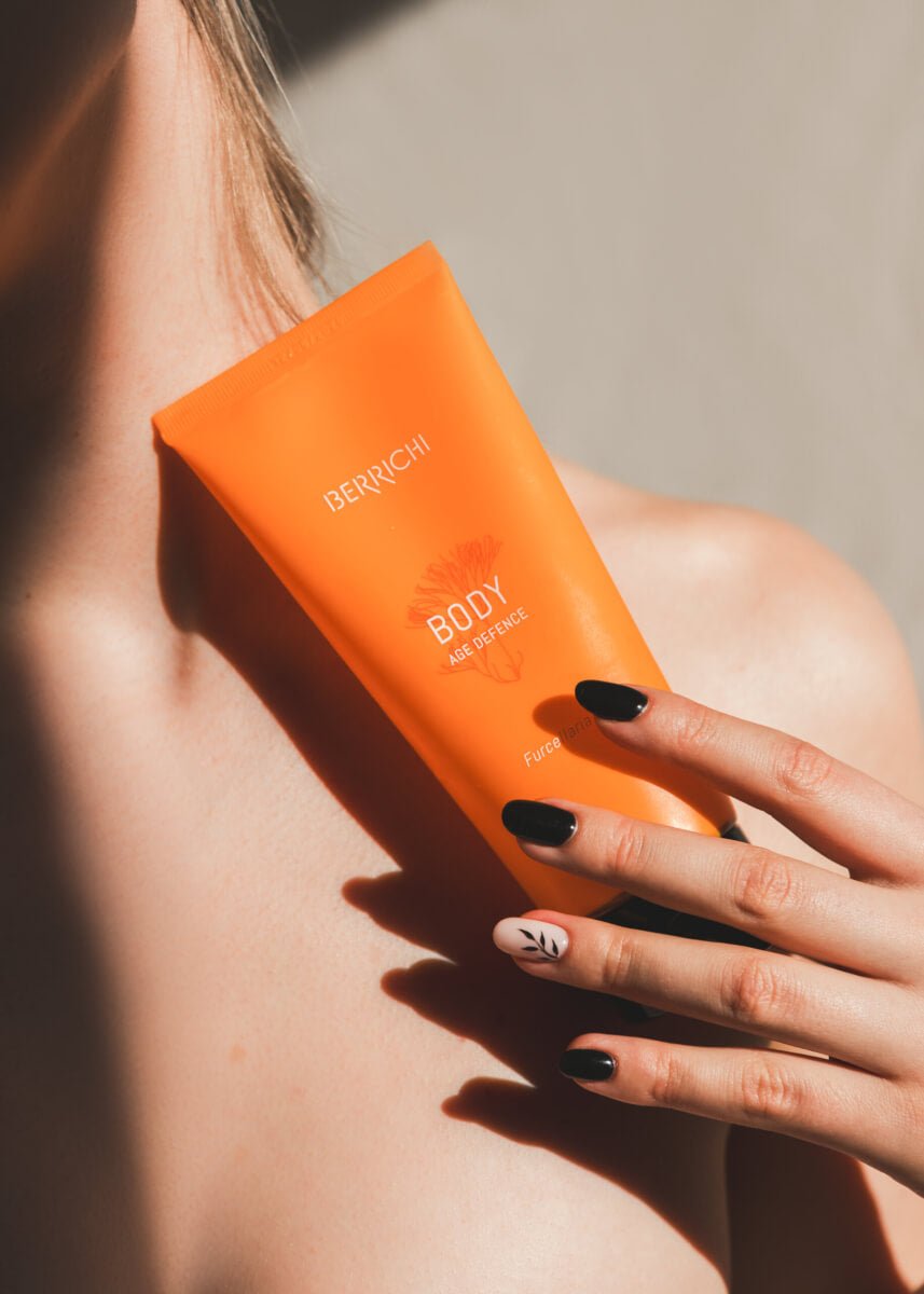 Orange krämtub med Age Defence Body cream från Berrichi hålls i händer med svarta naglar och neutral bakgrund