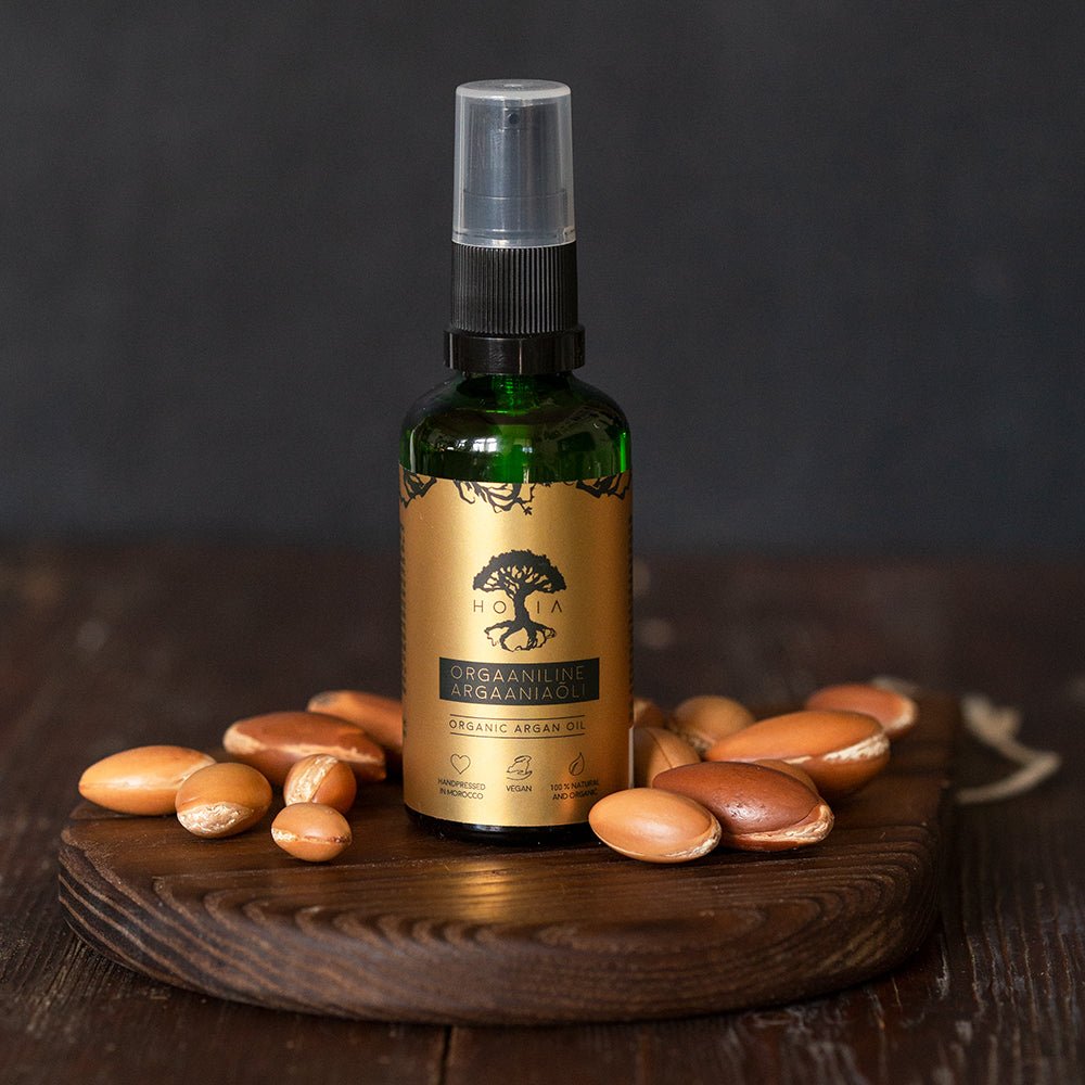 Organic Argan Oil 50ml i grön glasflaska med gyllene etikett och spraypump, omgiven av argannötter på träbricka