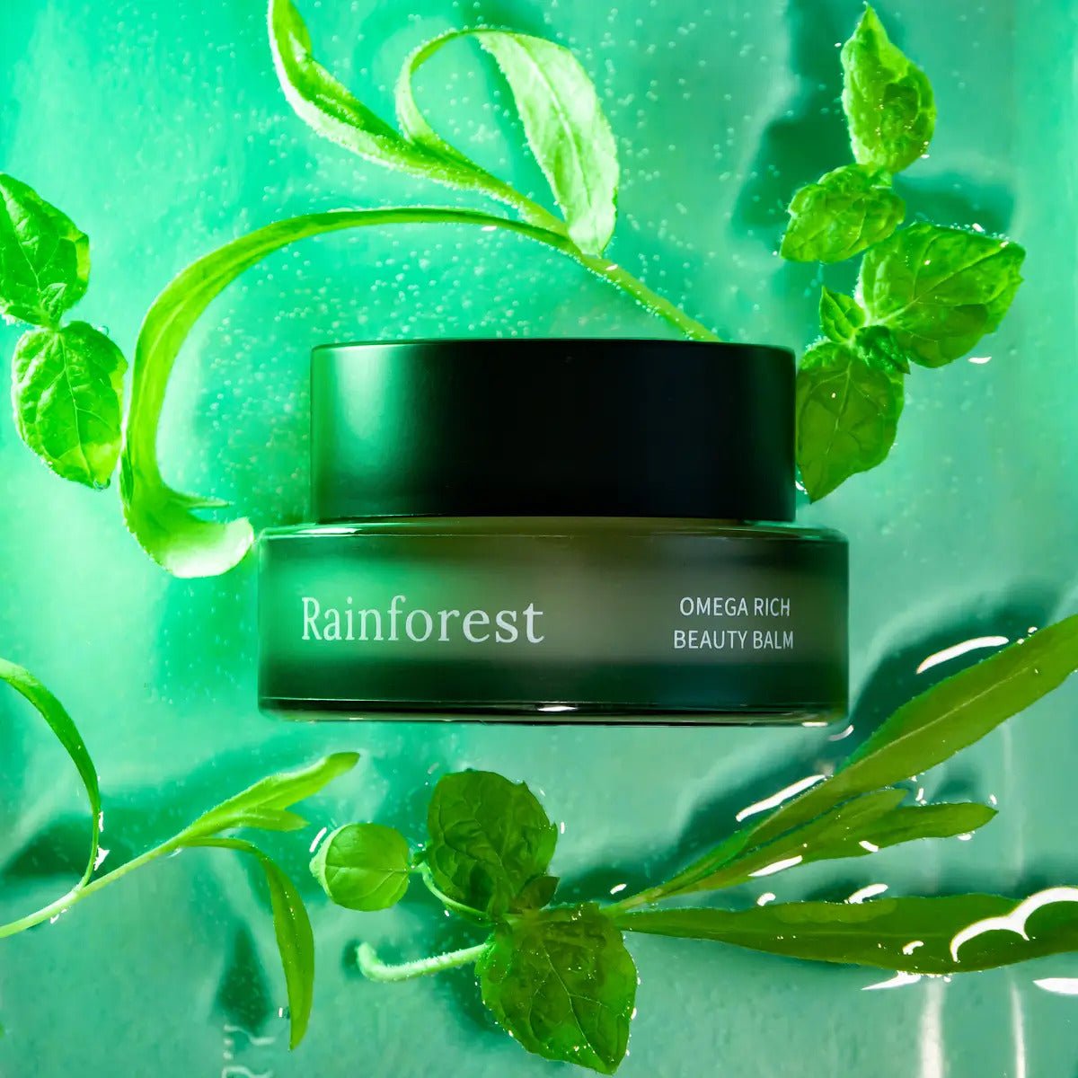 Rainforest Omega Rich Beauty Balm i grön glasburk med svart lock, omgiven av gröna växter och vattendroppar