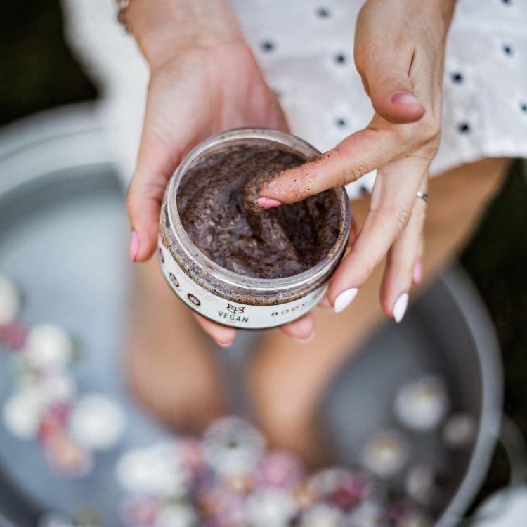 Händer håller Coffee Body Scrub 300g burk med brun kaffescrub, finger testar den grovkorniga texturen