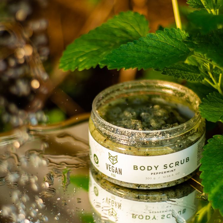 Öppen glasburk med grön Pepparmint Body Scrub omgiven av färska pepparmintblad på reflekterande yta