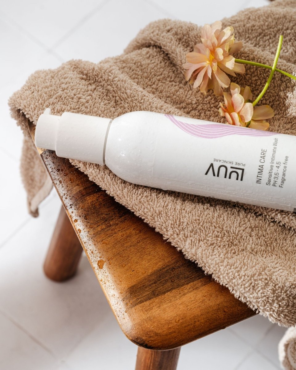Vit flaska med Sensitive Intimate Wash 200ml på beige handduk, dekorerad med blommor på träbänk